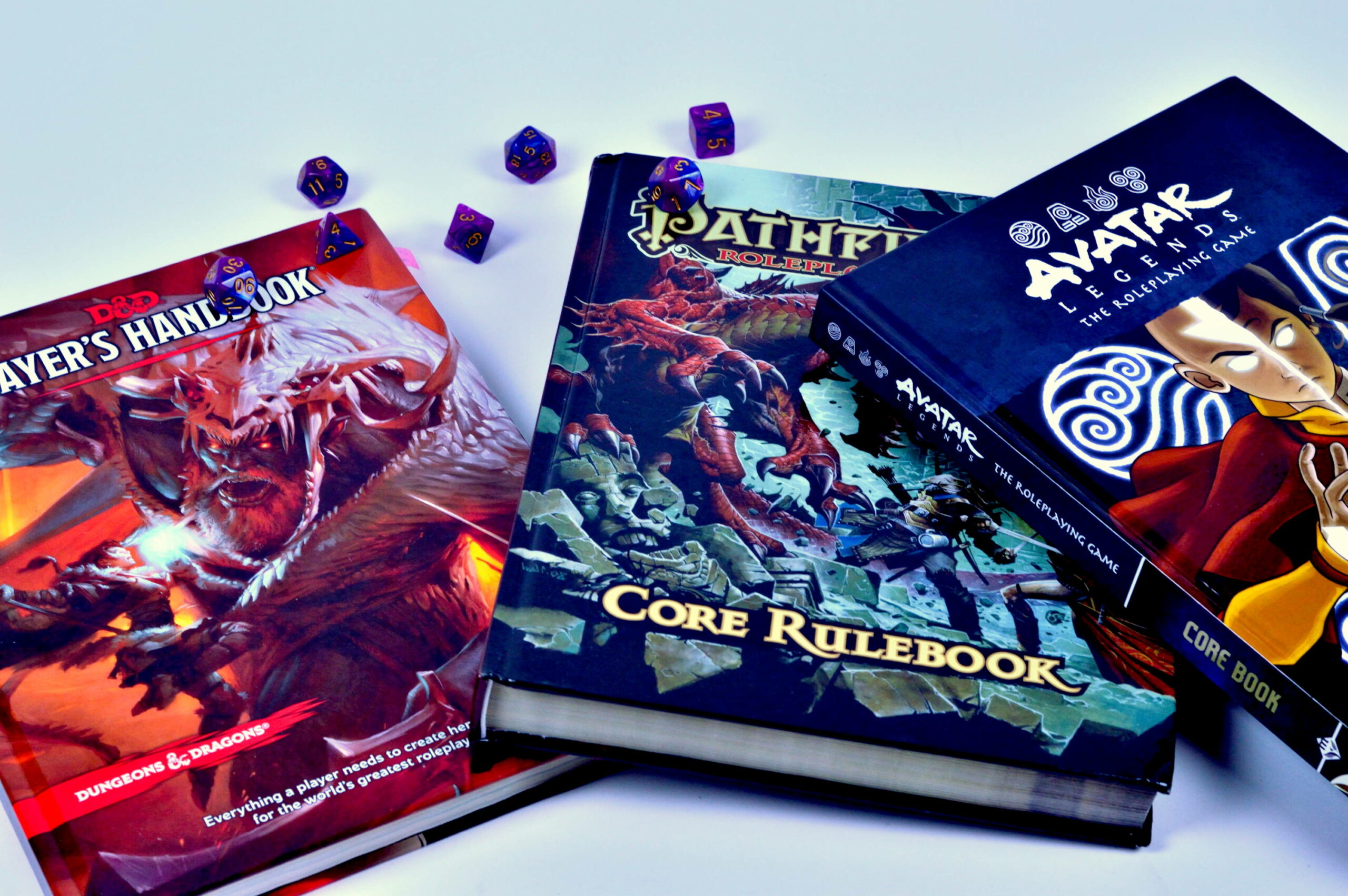 Tabletop Roleplaying Game rulebooks (D&D 5e, Pathfinder 1e, Avatar: The Last Airbender) and some purple dice.